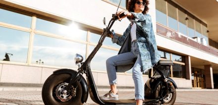 Full length image of pretty smiling curly woman in sunglasses sitting on modern motorbike outdoors and looking away