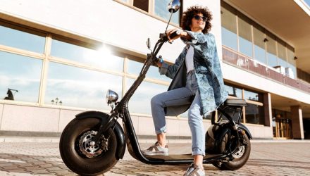 Full length image of pretty smiling curly woman in sunglasses sitting on modern motorbike outdoors and looking away