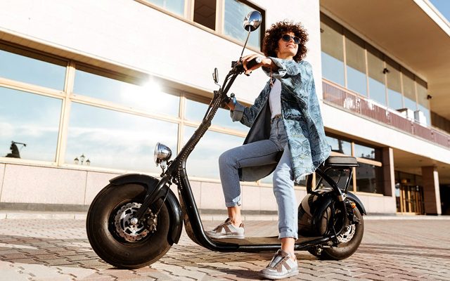 Full length image of pretty smiling curly woman in sunglasses sitting on modern motorbike outdoors and looking away